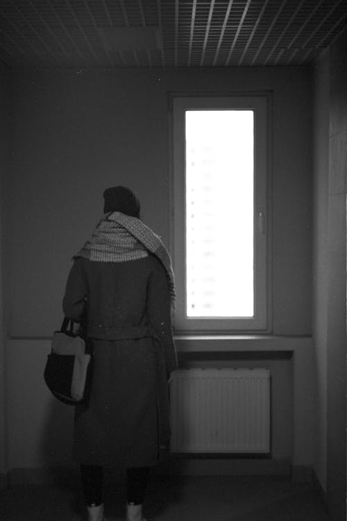 Grayscale Photo of a Person Standing Near a Window