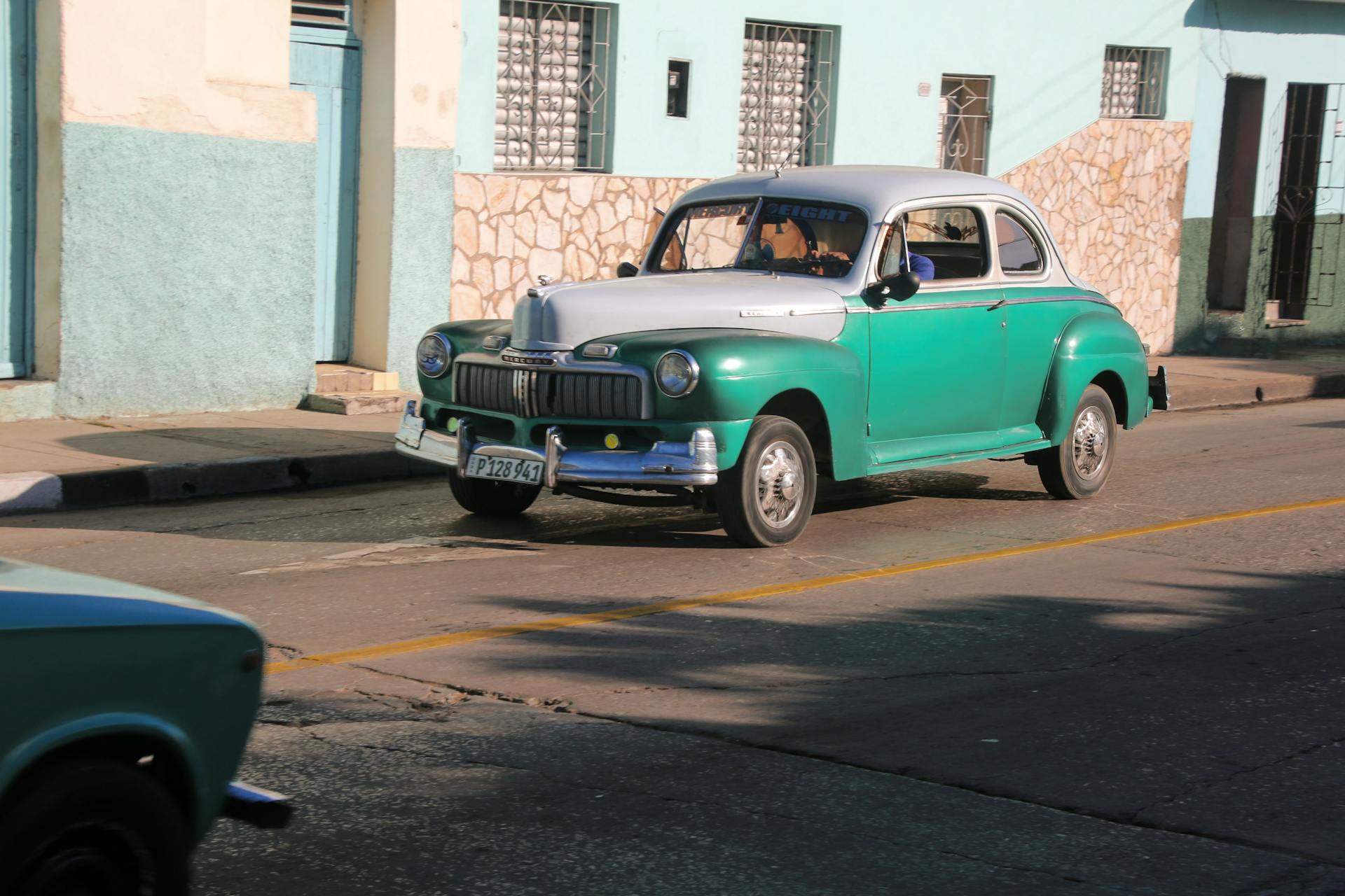 Classic Mercury Eight automobile driving on a sunny street with pastel buildings.