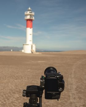A DSLR camera on a tripod capturing a scenic lighthouse on a sandy beach under a clear sky.