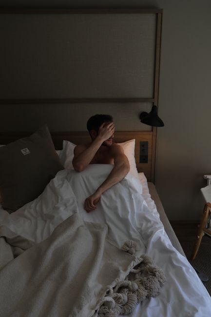 A shirtless man lounging in bed, covering his face in a cozy bedroom in Zermatt.