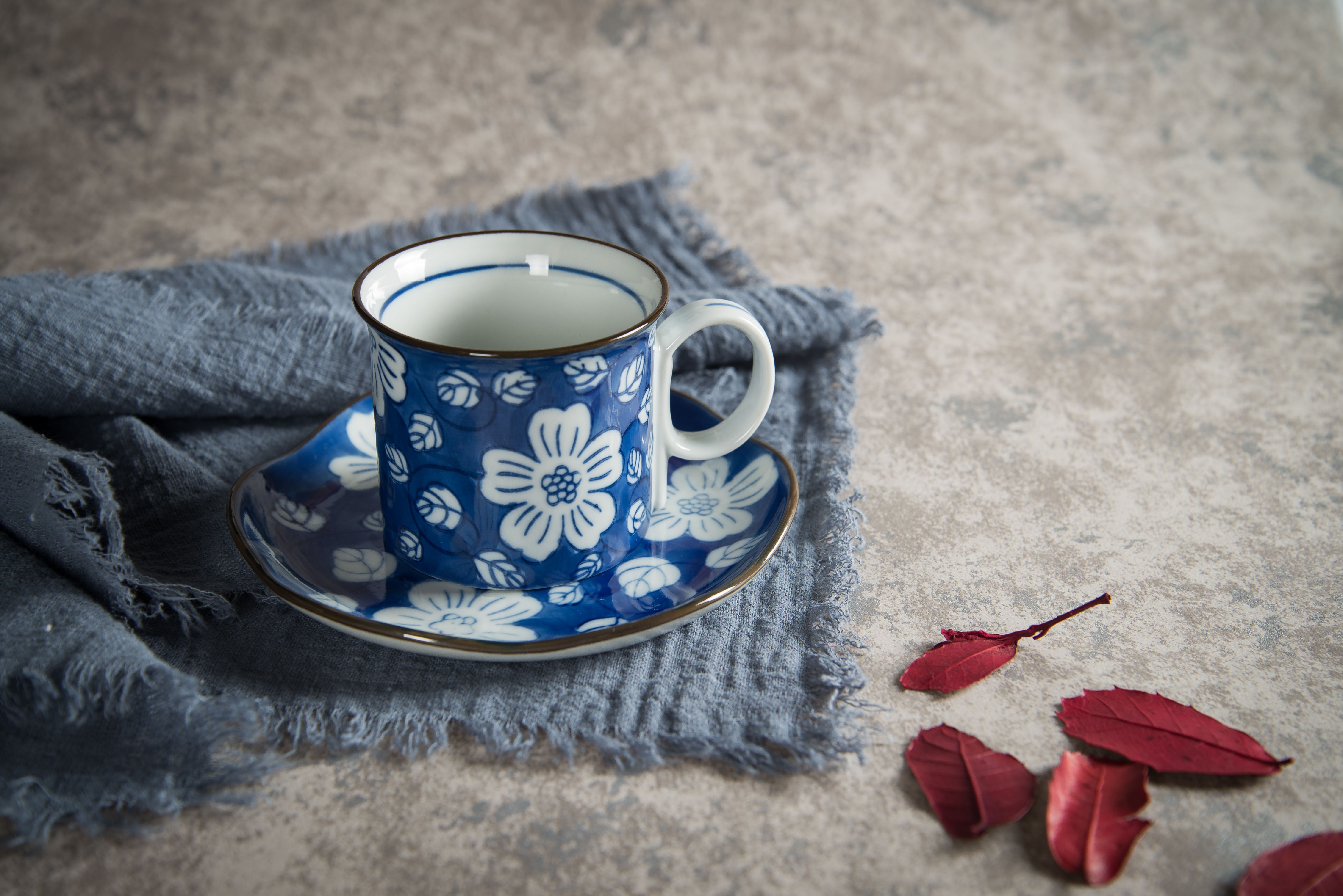 Elegant floral porcelain coffee mug on a blue cloth with red leaves and textured background.