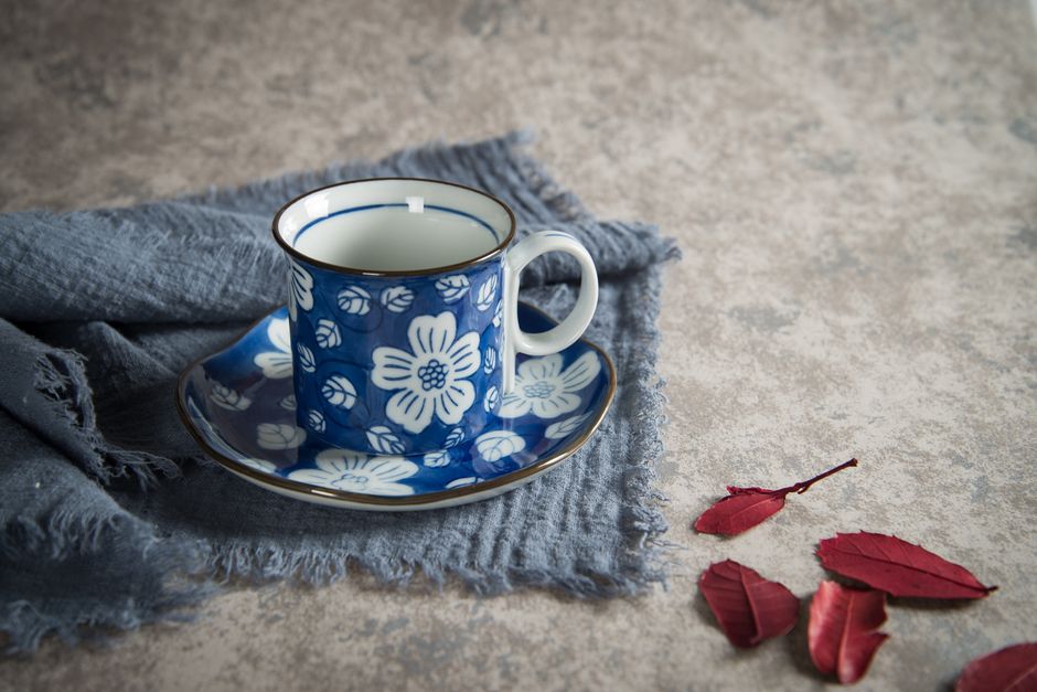 Elegant floral porcelain coffee mug on a blue cloth with red leaves and textured background.