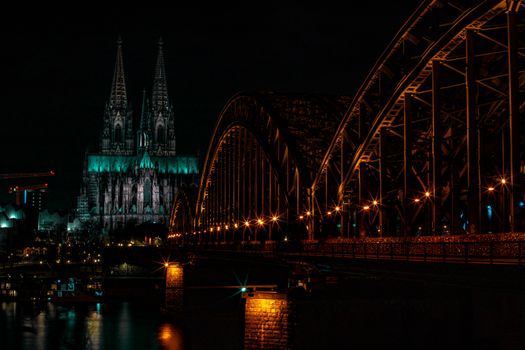 Illuminated view of Cologne Cathedral and Hohenzollern Bridge spanning the Rhine River at night.