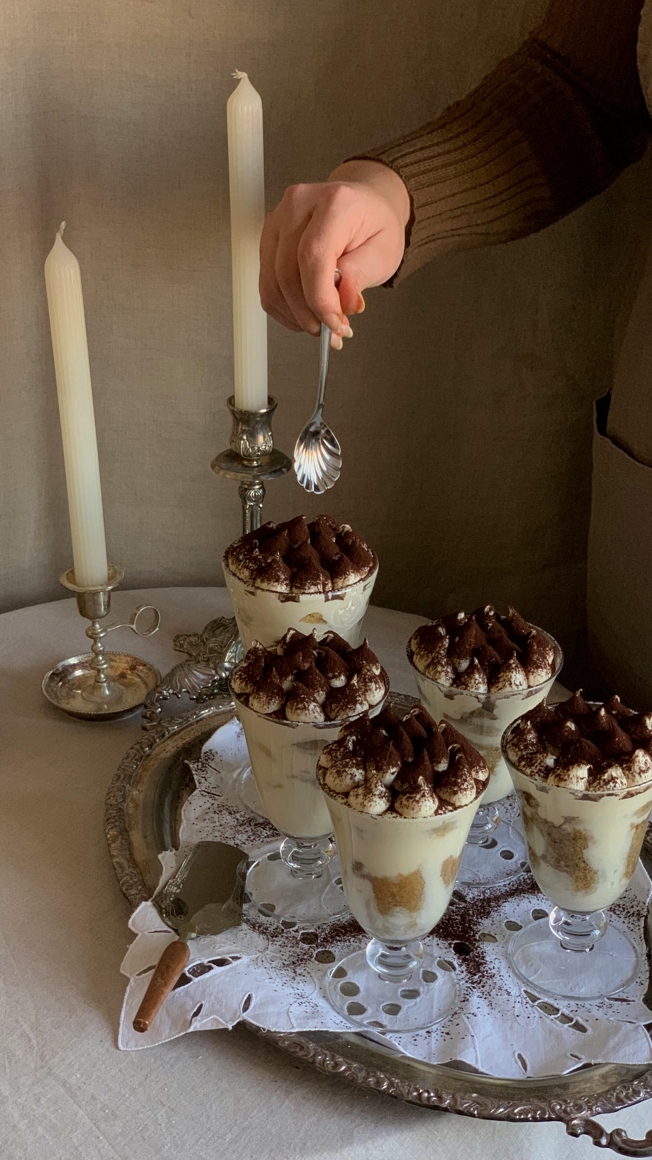 A hand garnishes luxurious tiramisu desserts in elegant glass cups, set on a vintage tray with candles.