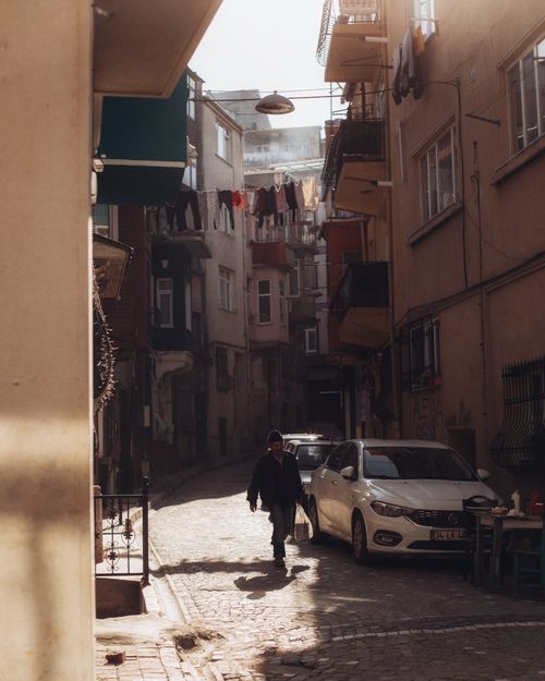 Free A man walking in a sunlit urban alley with parked cars and hanging laundry, capturing the essence of daily city life. Stock Photo