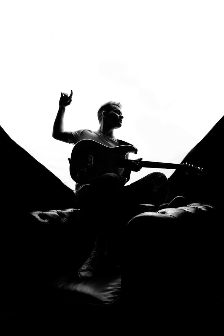 Black And White Picture Of Man Sitting On A Leather Armchair Playing A Guitar