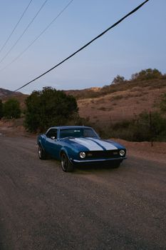Classic blue Camaro on a scenic country road during sunset, showcasing automotive nostalgia.