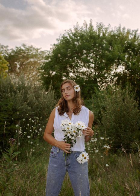 A woman standing in a meadow holding a bouquet of daisies, with flowers in her hair, during sunset.