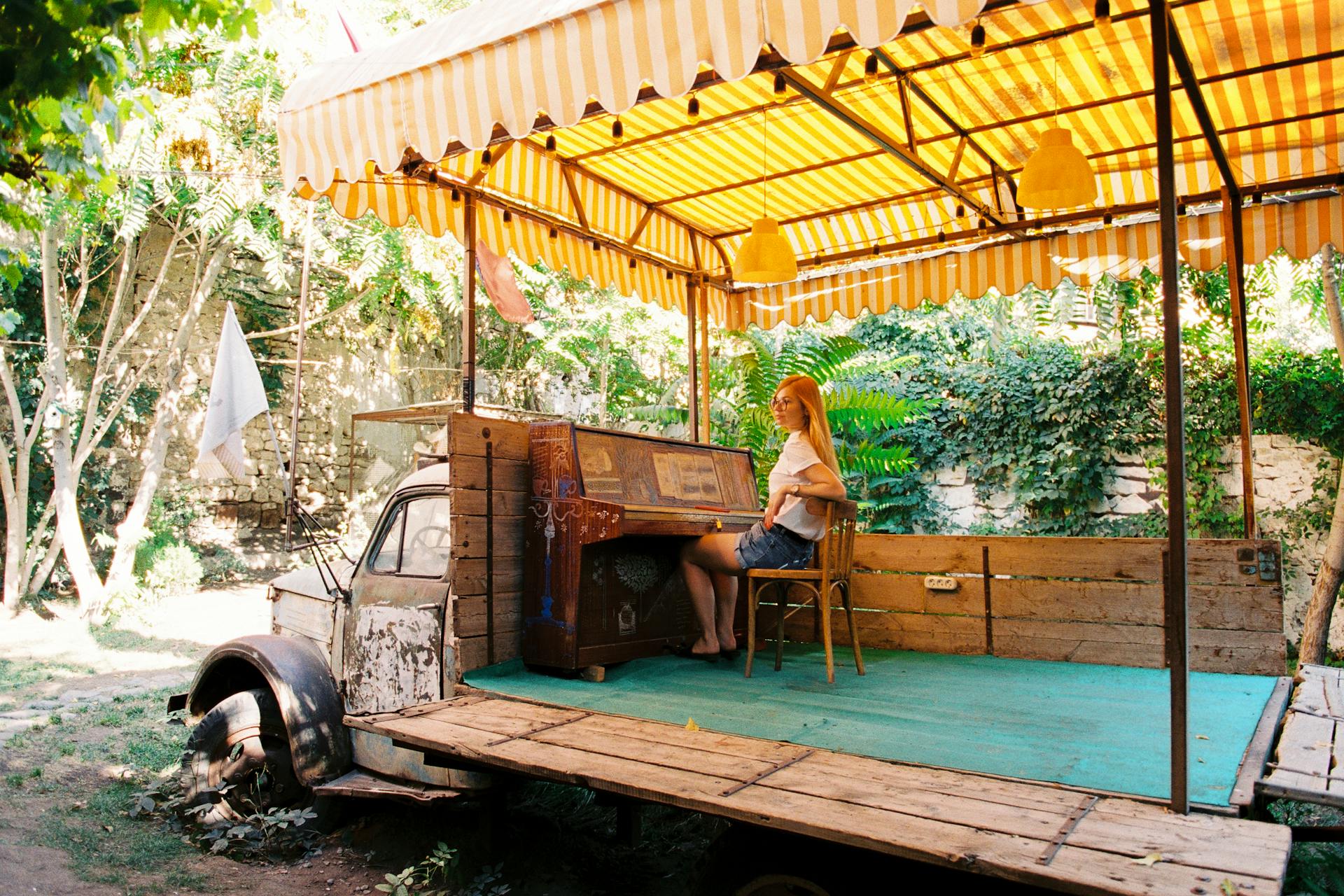 A woman sits at a piano on a vintage truck under a striped canopy in a lush backyard setting.