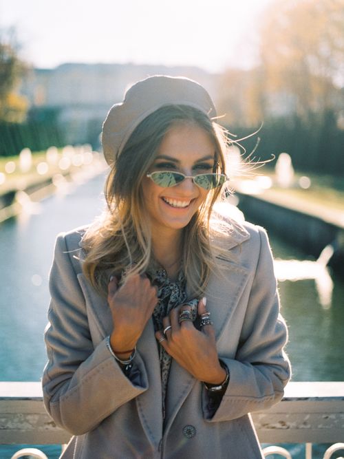 Free A fashionable woman wearing sunglasses and a coat smiles in a sunny park setting. Stock Photo
