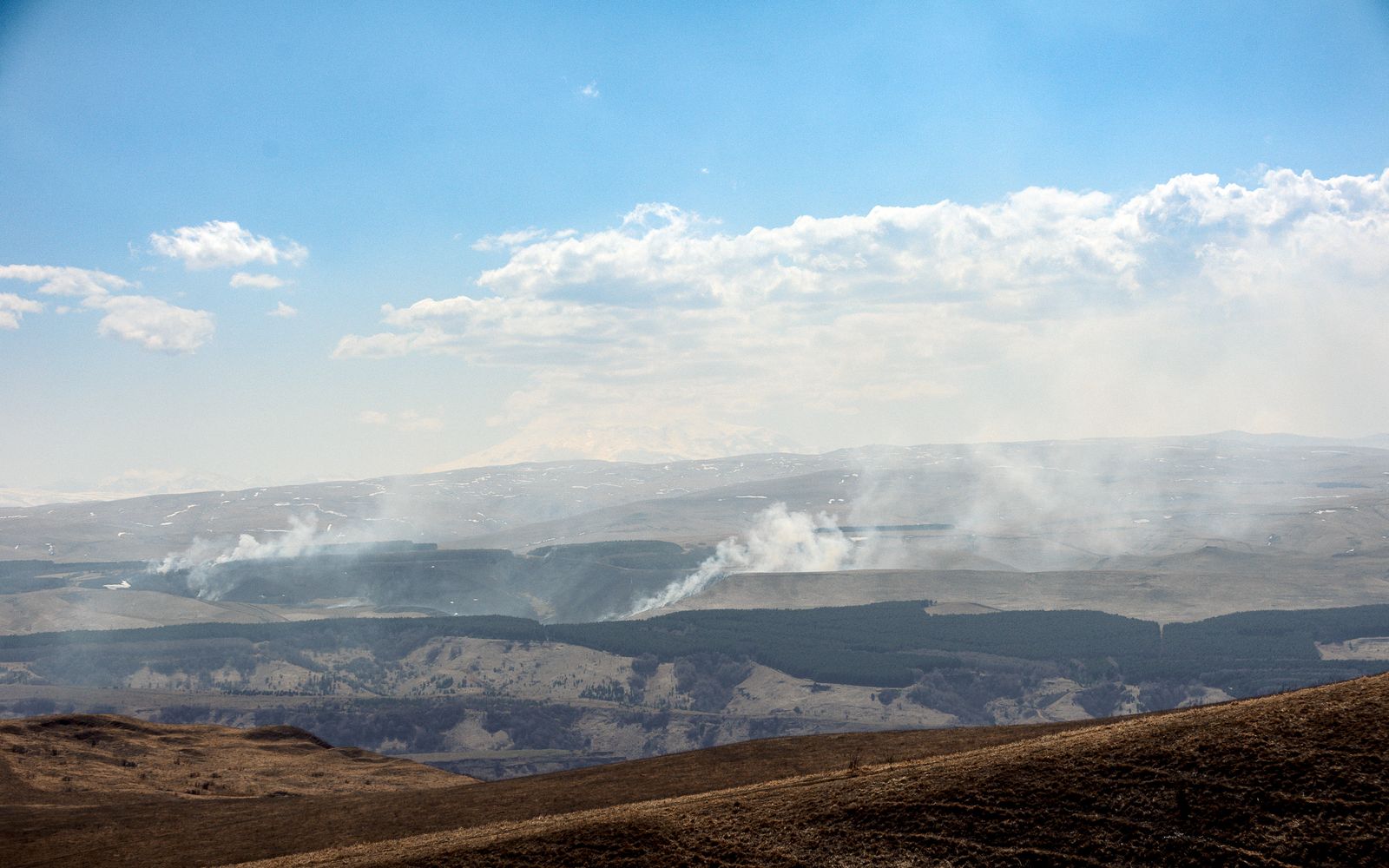 Smoke plumes rising from a forested valley in a hilly landscape under a partly cloudy sky