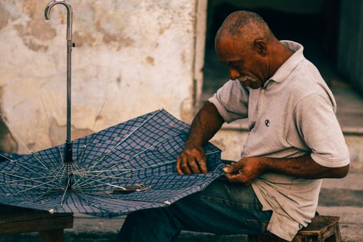An elderly man carefully repairs an umbrella in a rustic outdoor setting, showcasing craftsmanship and focus.