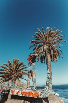 Tattooed woman standing by palm trees and ocean on a sunny day, embracing summer vibes.