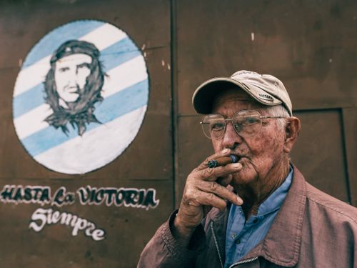 Free Elderly man with eyeglasses smokes a cigar outdoors, standing by a Che Guevara mural. Stock Photo
