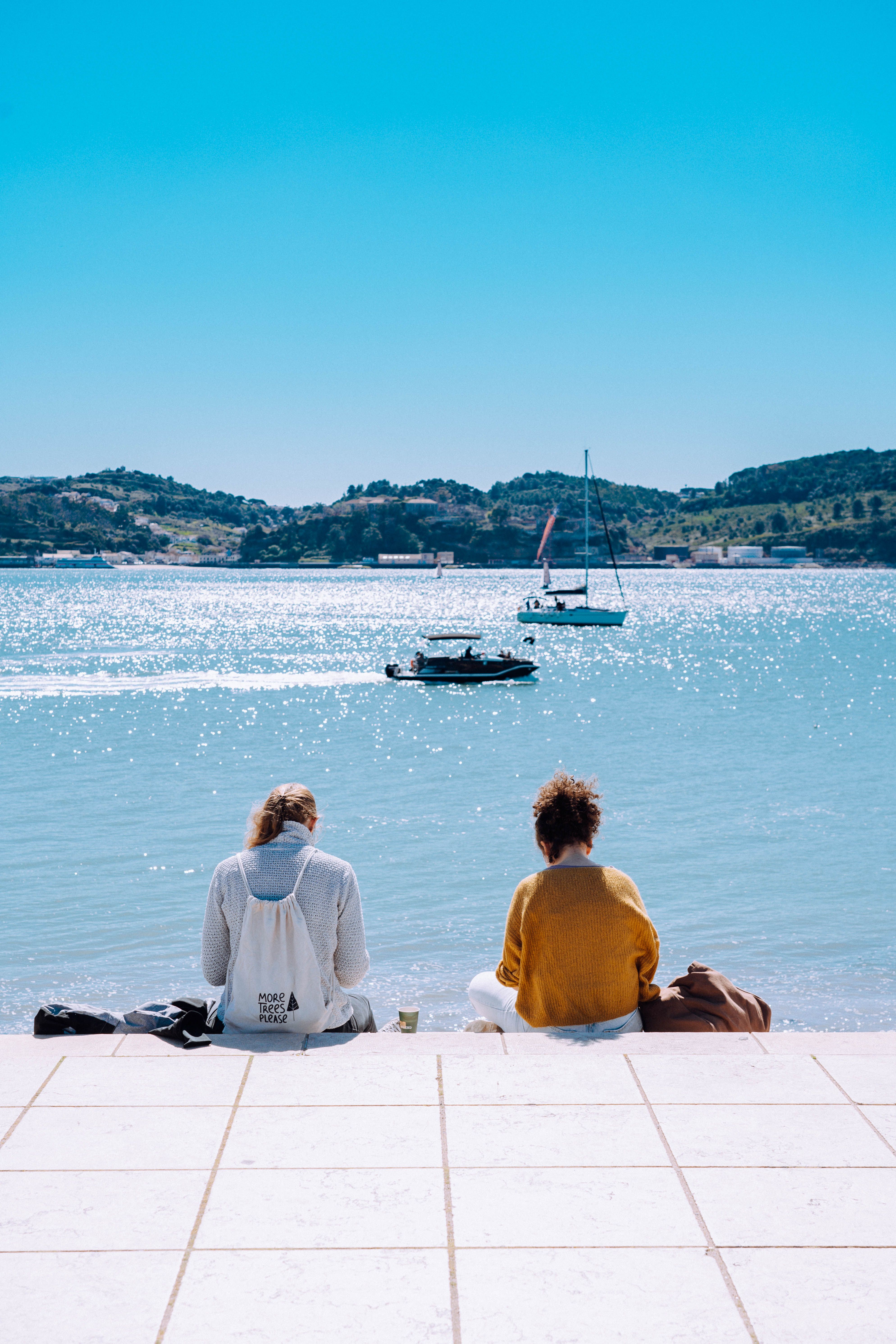 Two women sit by the ocean in Lisbon, enjoying a sunny day with boats in view.