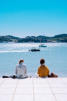 Two women sit by the ocean in Lisbon, enjoying a sunny day with boats in view.