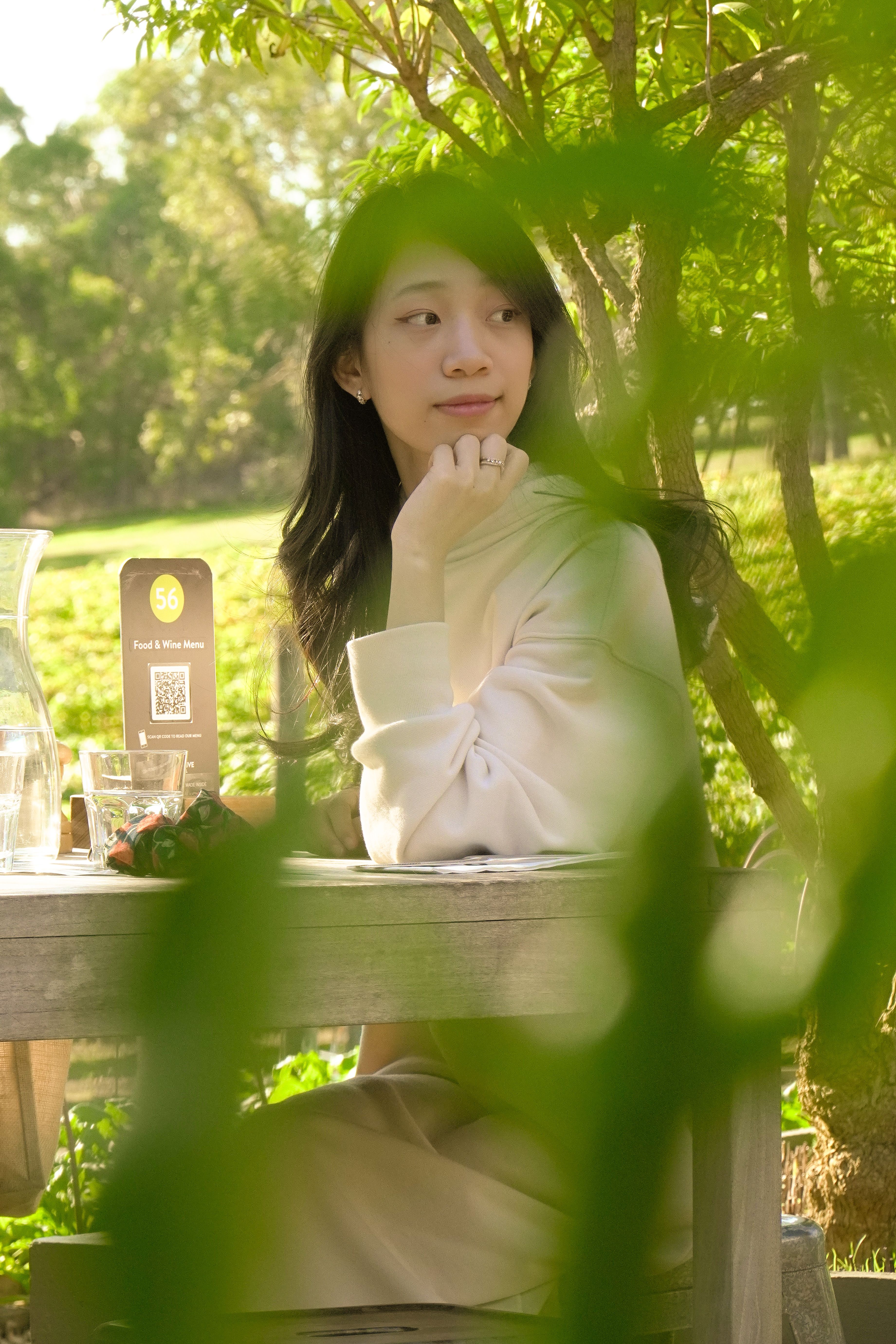 Asian woman sitting thoughtfully at a table, surrounded by greenery in a sunny outdoor setting.