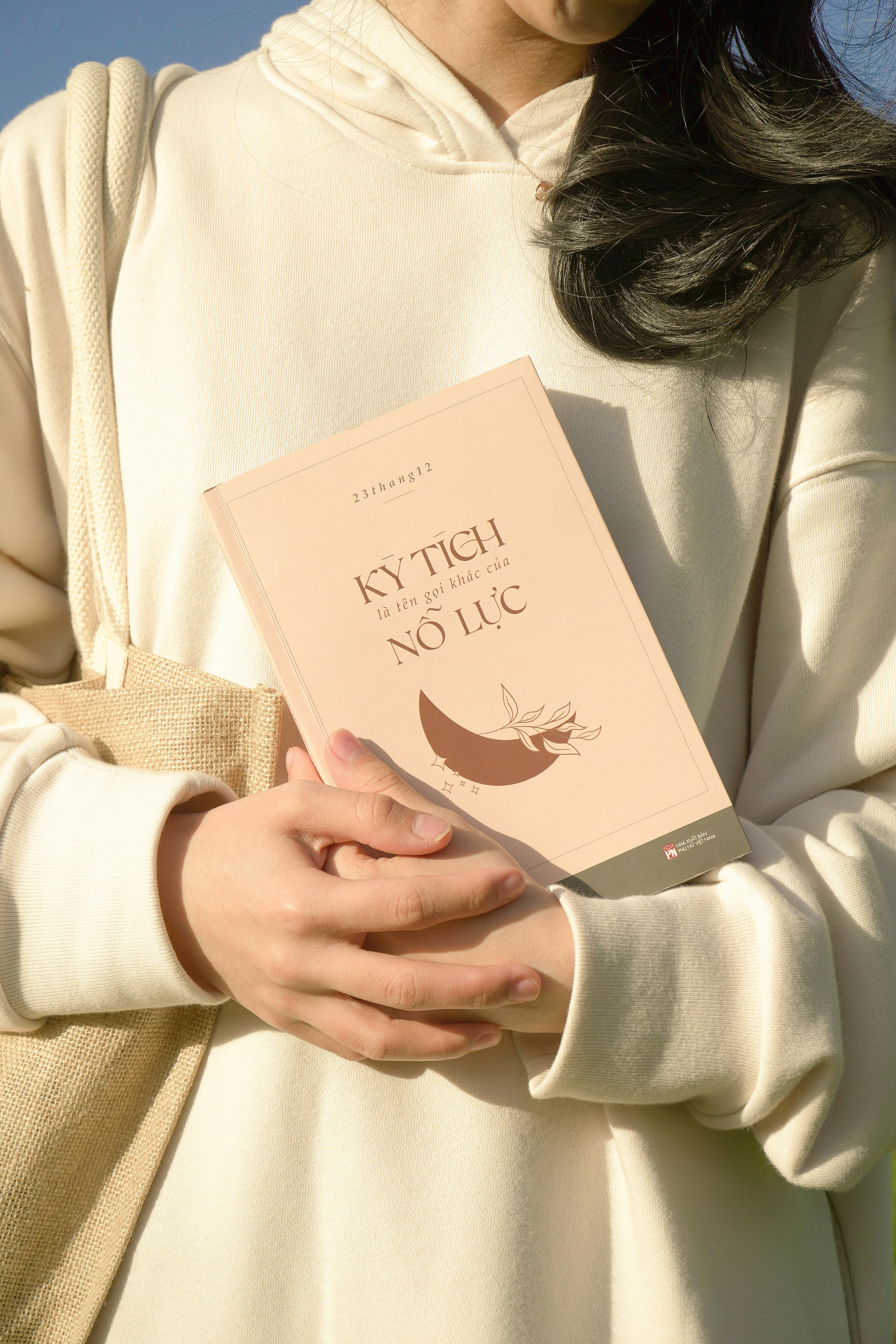 Person in white hoodie holding a book outdoors, capturing a serene moment.