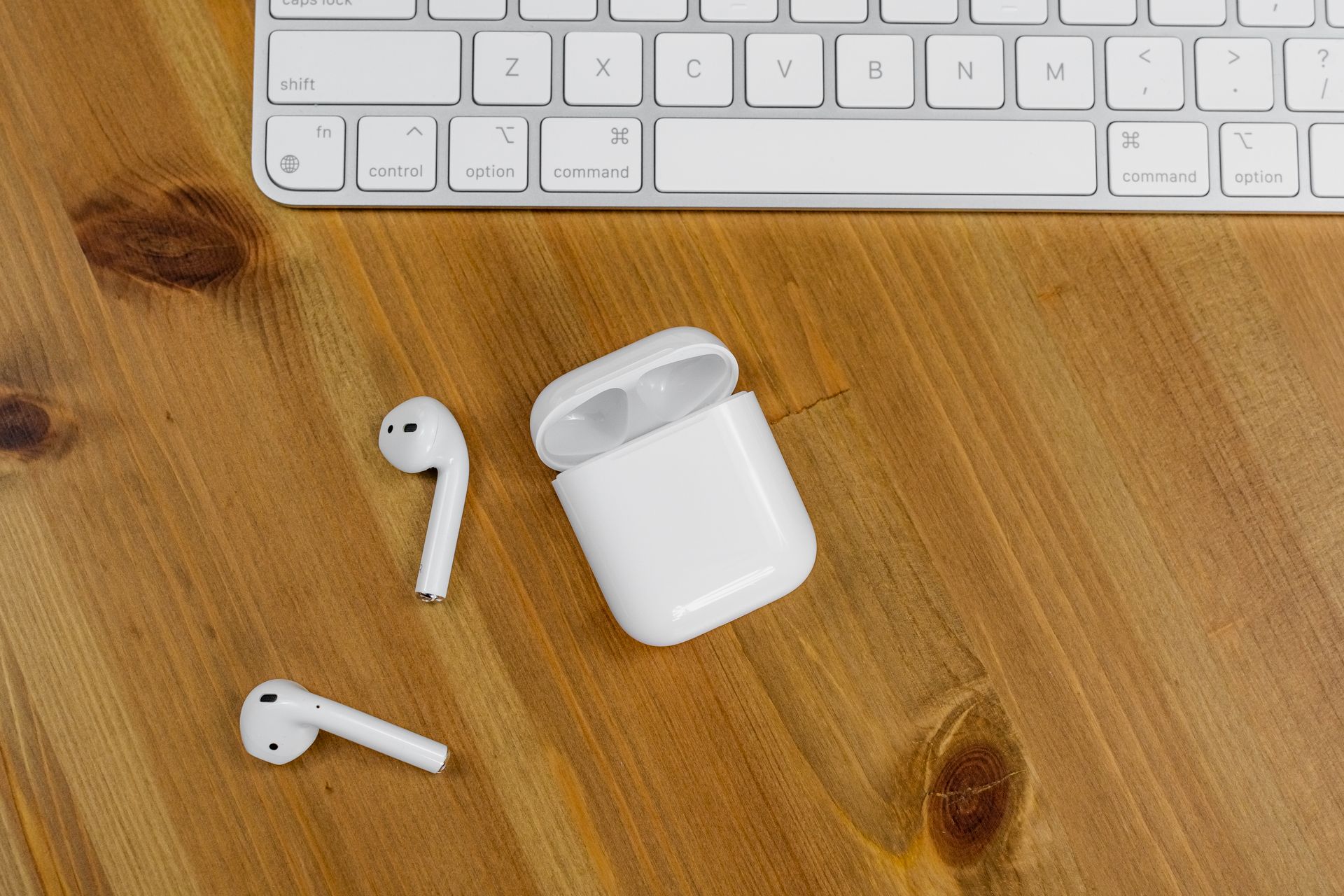 Close-up view of wireless earbuds next to an Apple keyboard on a wooden desk surface.
