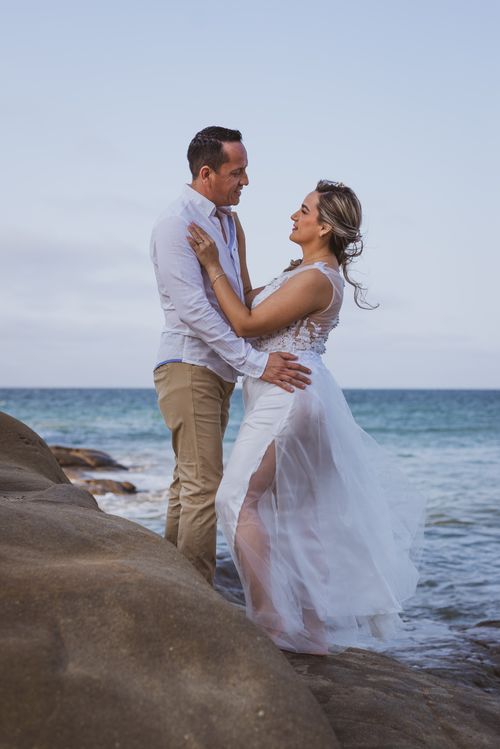 Free Couple embracing on rocky beach in wedding attire, capturing a romantic moment by the sea. Stock Photo