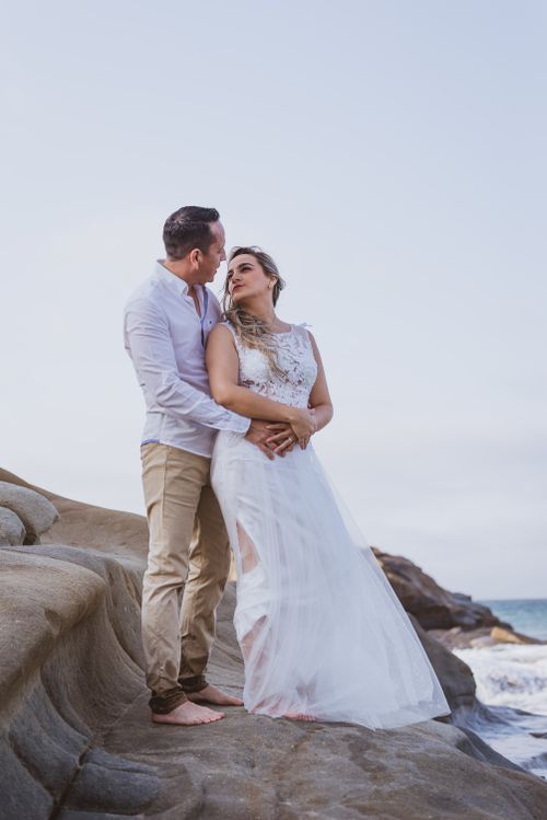 Free A couple embraces on coastal rocks in wedding attire during sunset. Stock Photo