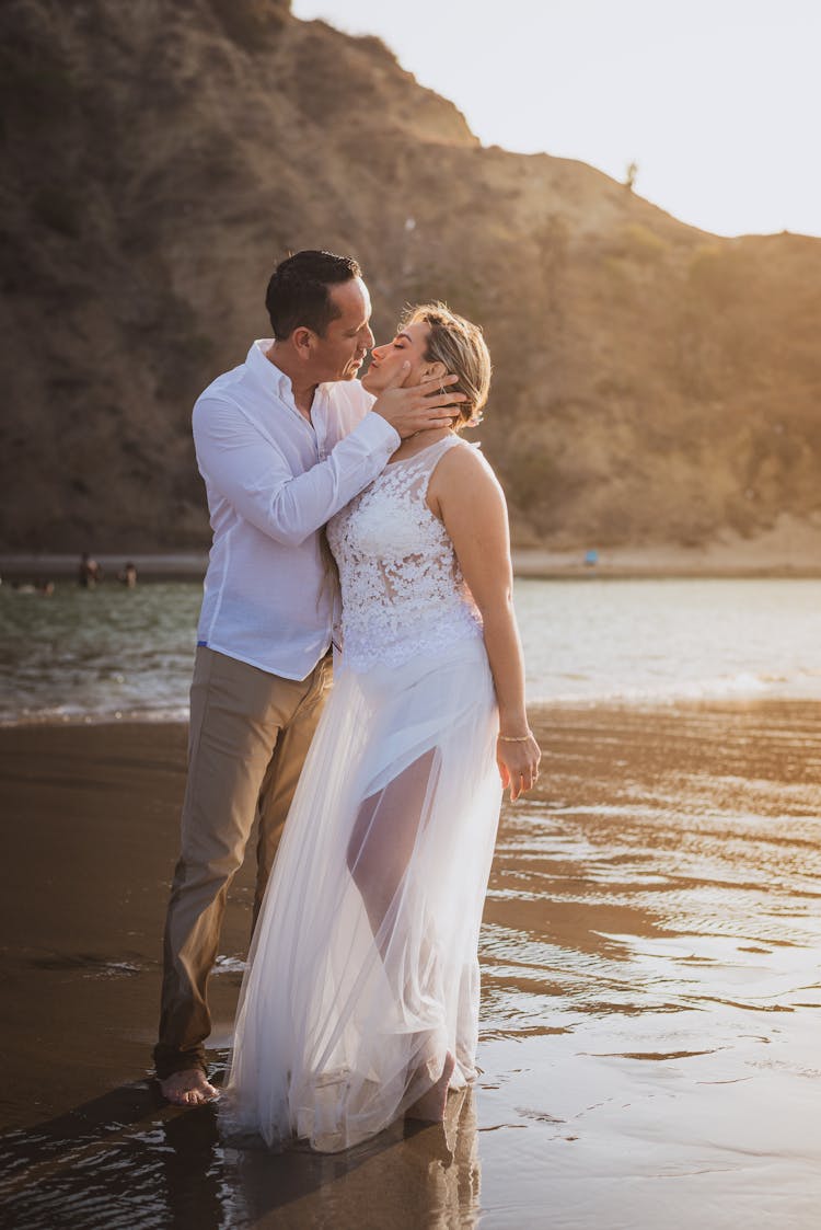 A Man Kissing A Woman In A White Dress At The Beach