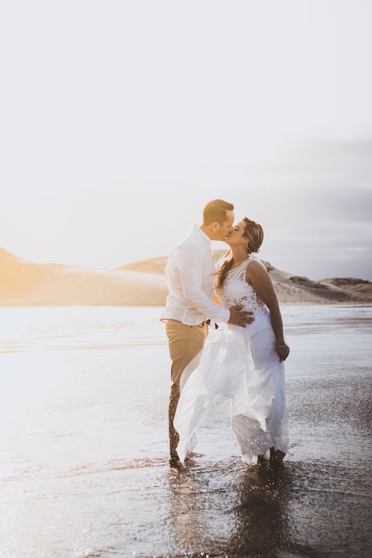 A Woman In A White Dress Kissing A Man At The Beach