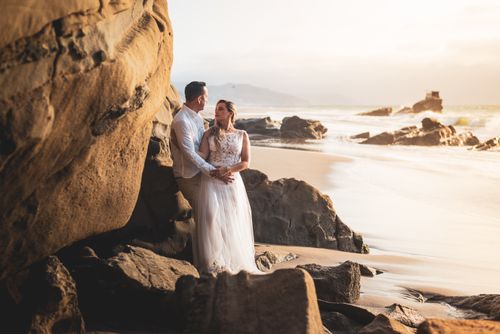 Free A couple shares a romantic moment during their wedding photoshoot on a scenic beach at sunset. Stock Photo