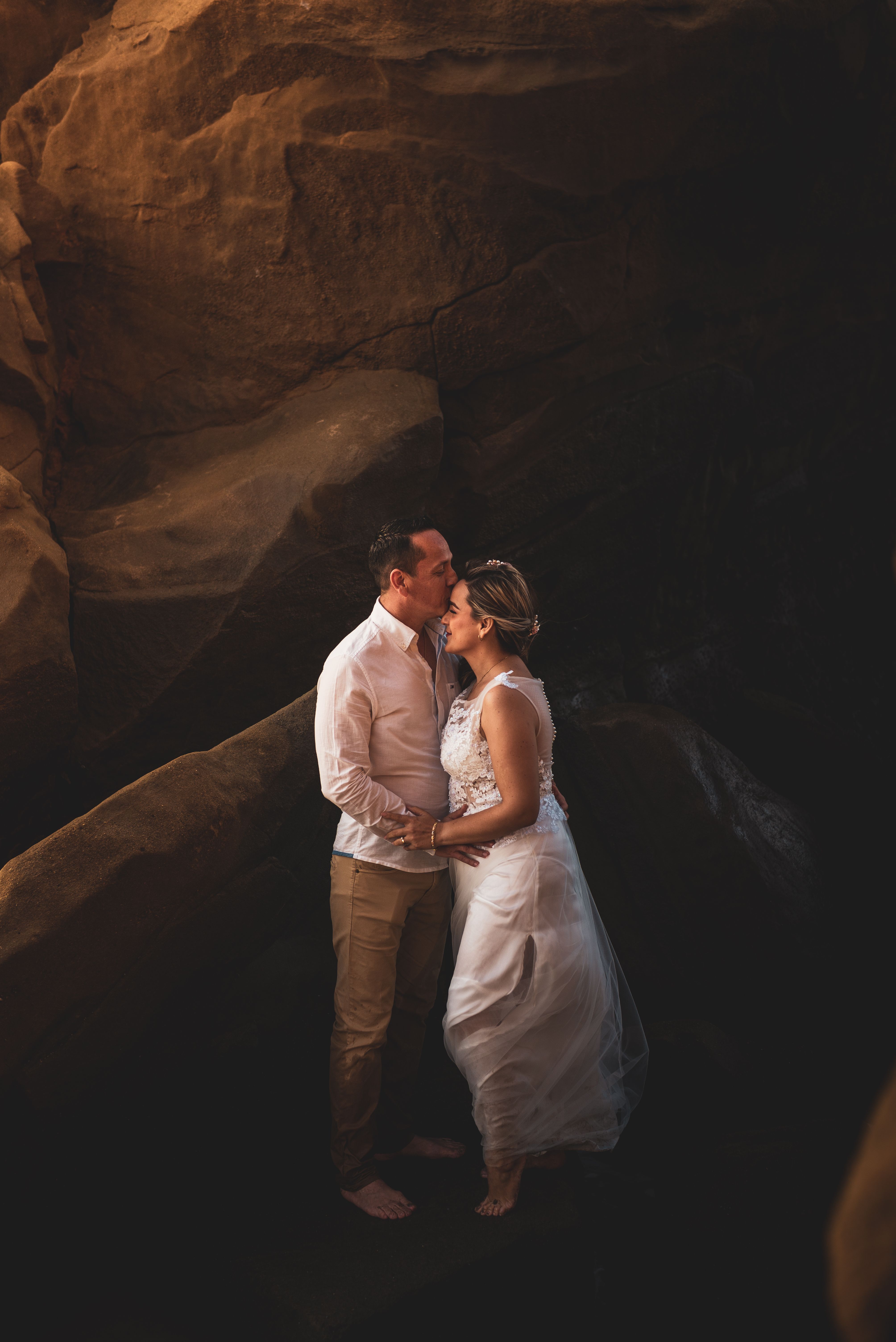 A couple shares a romantic moment kissing near rocky formations by the beach, conveying love and intimacy.