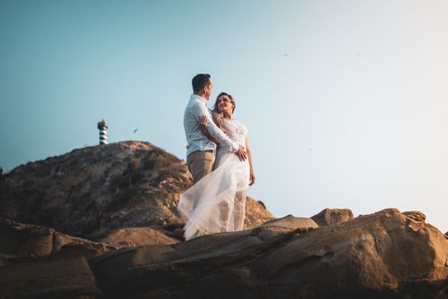 Free Newlyweds embracing on rocky terrain with a lighthouse in the background. Dreamy outdoor photography. Stock Photo