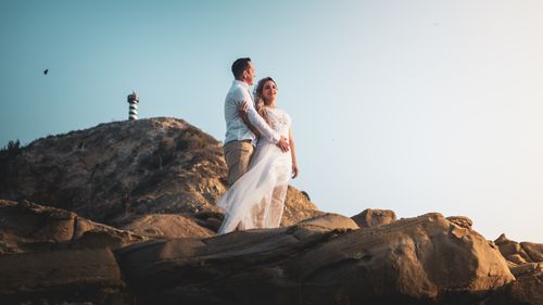 Free A couple embraces on rocky cliffs with a lighthouse in the background. Perfect wedding capture. Stock Photo