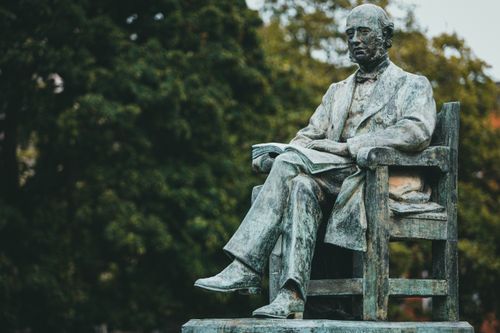 Free Elegant bronze statue of a man seated in a chair surrounded by lush greenery. Stock Photo