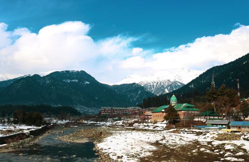 Free Picturesque snowy mountains with a flowing river and green-roofed buildings in a rural valley. Stock Photo
