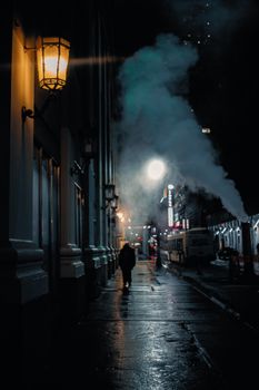 Atmospheric night view of a city street with steam and lamps illuminating a pedestrian.
