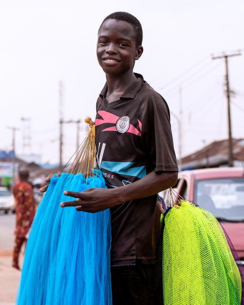 Free A young Nigerian vendor smiling while selling colorful nets on a busy street. Stock Photo
