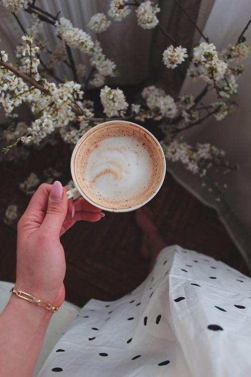 Free A stylishly dressed hand holds a cappuccino against a backdrop of spring blossoms. Stock Photo