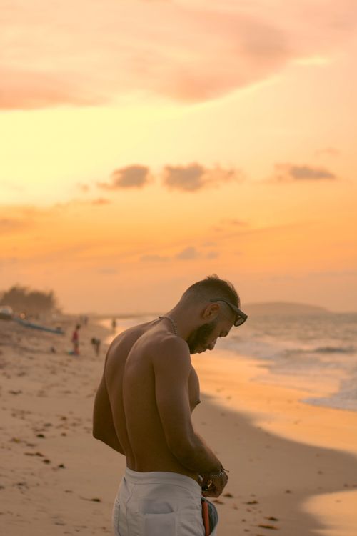 Free A contemplative man stands shirtless on a sandy beach during a beautiful sunset. Stock Photo
