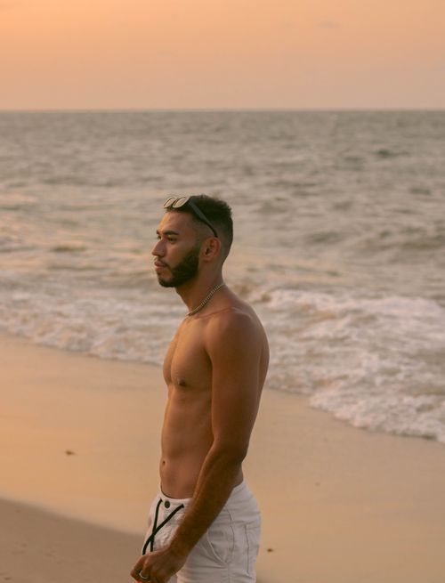 Free Shirtless man stands by the shore as the sun sets over the ocean. Stock Photo