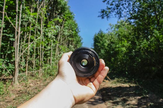 A hand holding a camera lens in a lush green forest under the bright daylight.