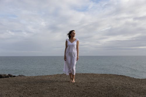 Photo of a Woman in a White Dress Standing Near the Ocean