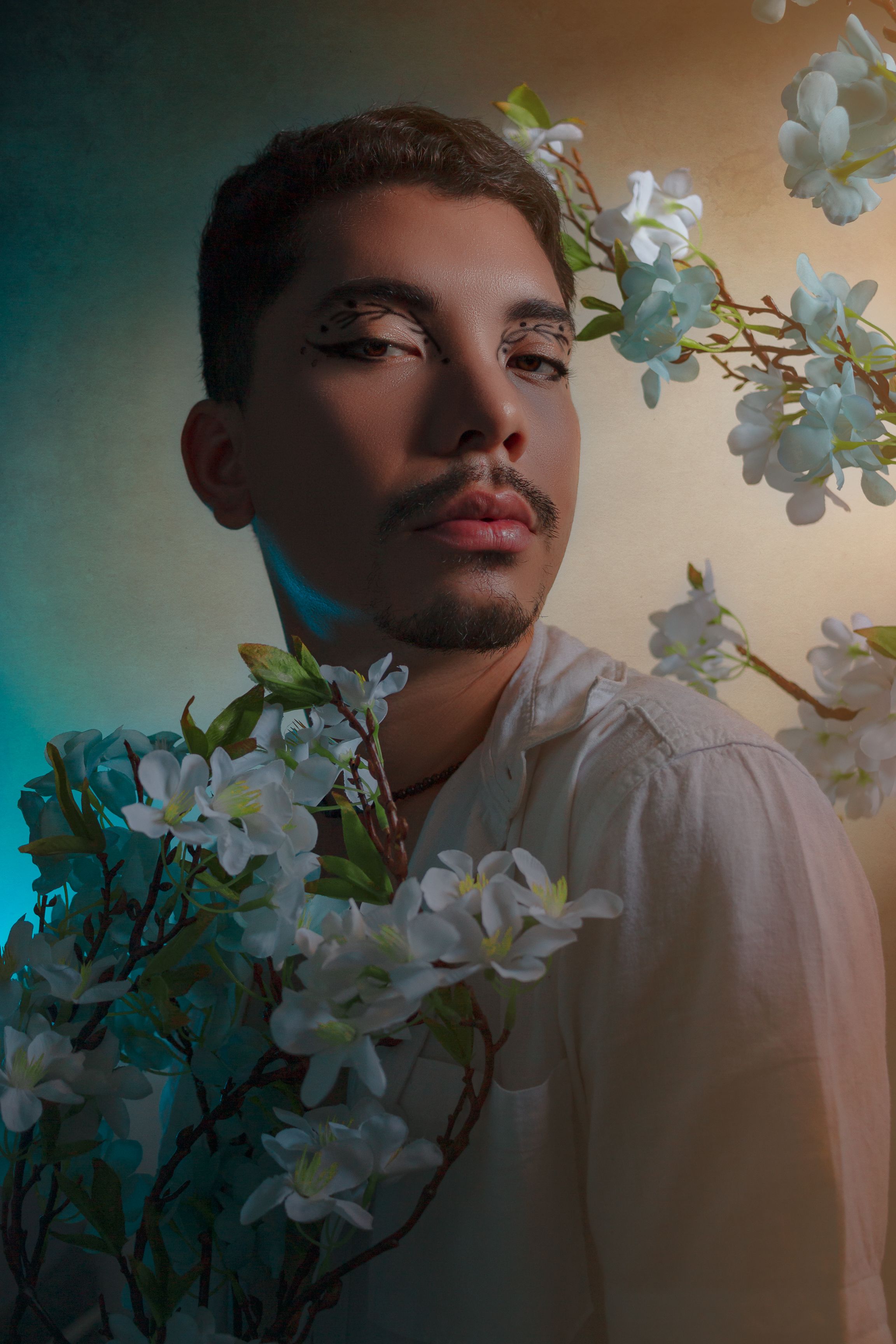 A creative portrait of a man with artistic makeup surrounded by blossoming jasmine flowers in a studio setting.