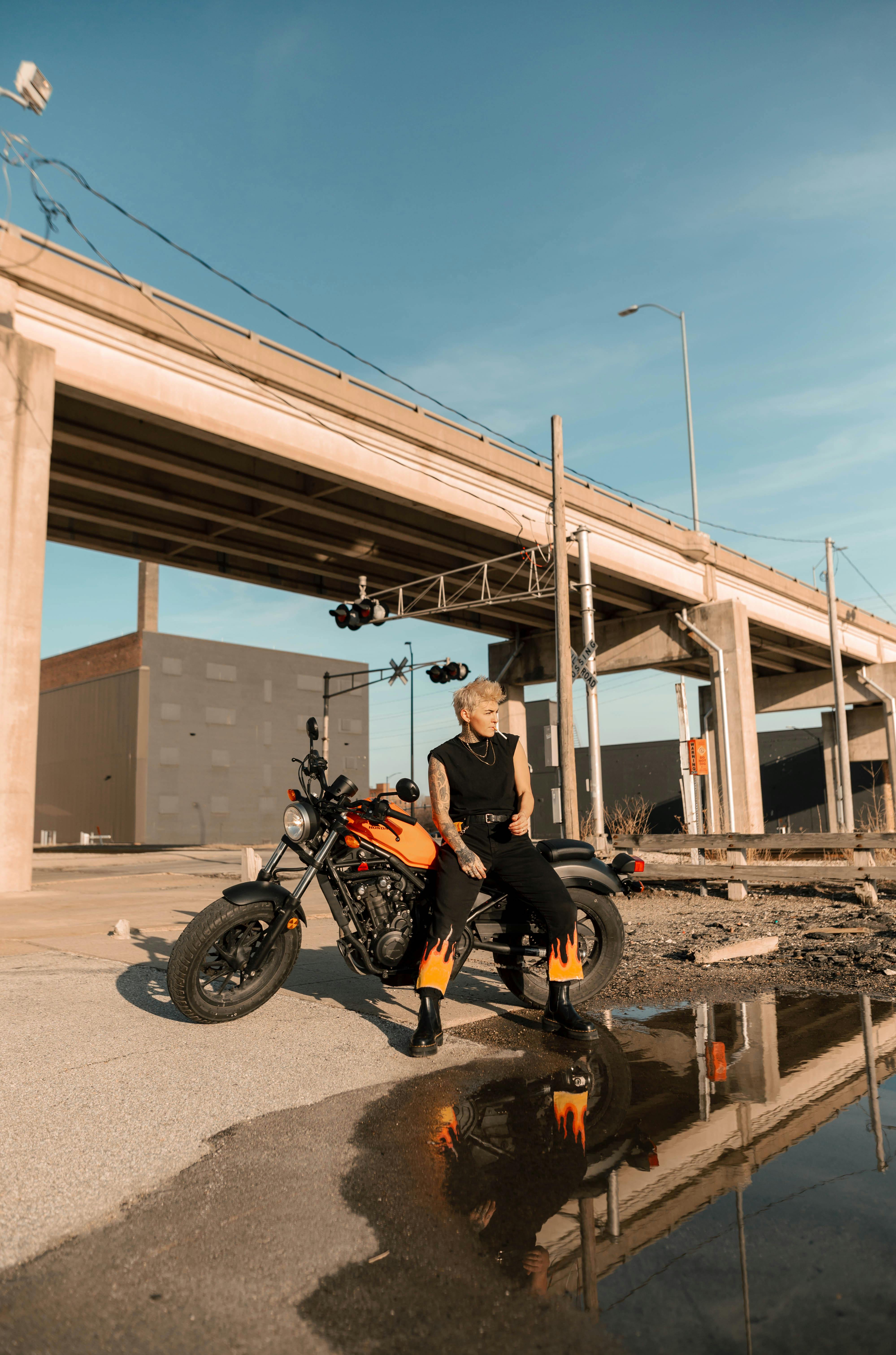 A fashionable person poses with a motorcycle under an urban bridge in daylight.