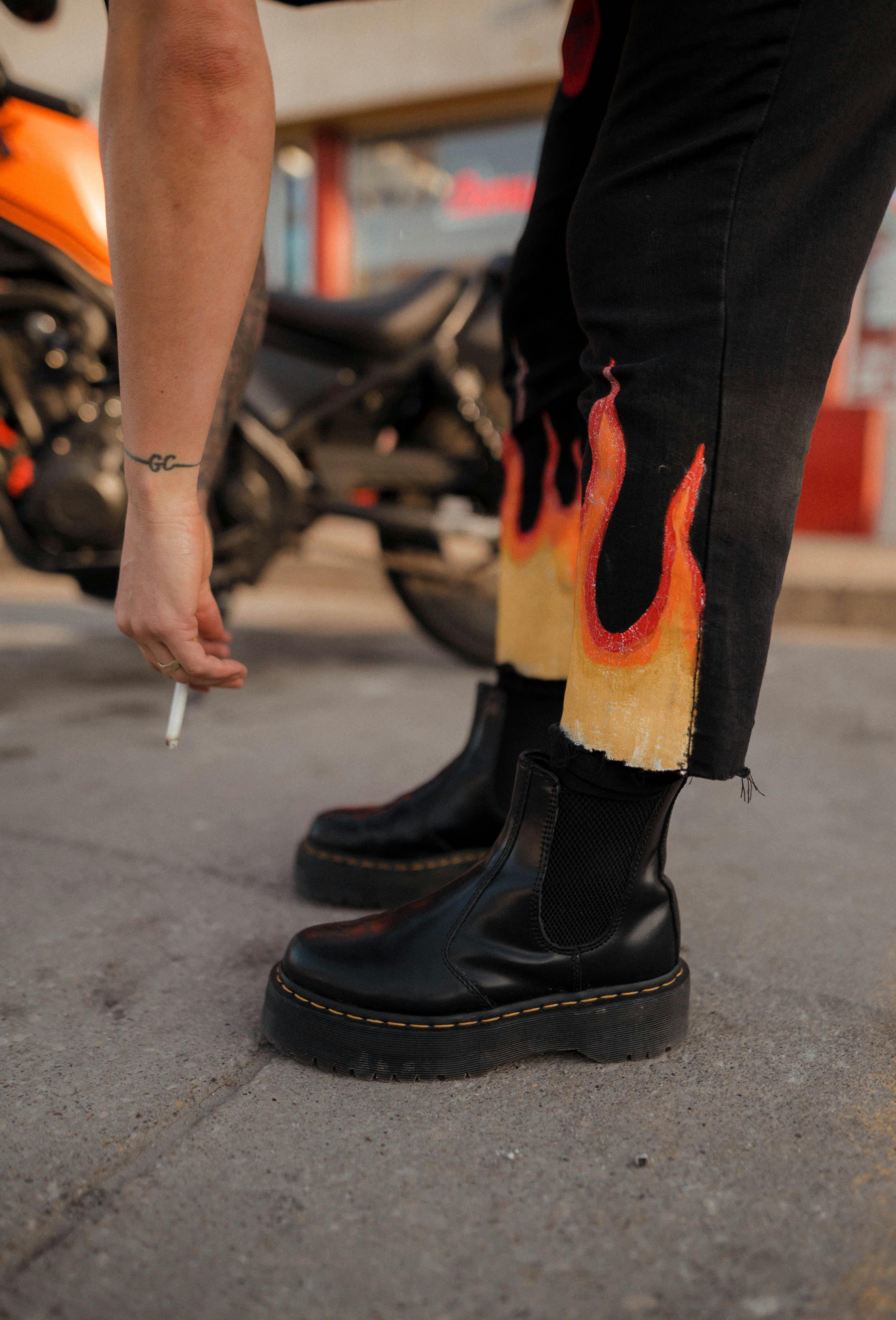 Close-up of trendy black boots paired with flame-patterned pants, featuring a motorbike in the background and a hand holding a cigarette.