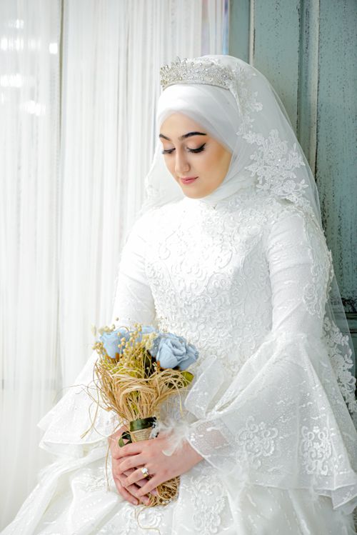 Free Stunning bride in a white lace gown and hijab, holding a delicate bouquet indoors. Stock Photo