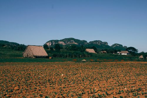 Free A picturesque rural farm featuring fields and thatched roofs against mountains under a clear summer sky. Stock Photo