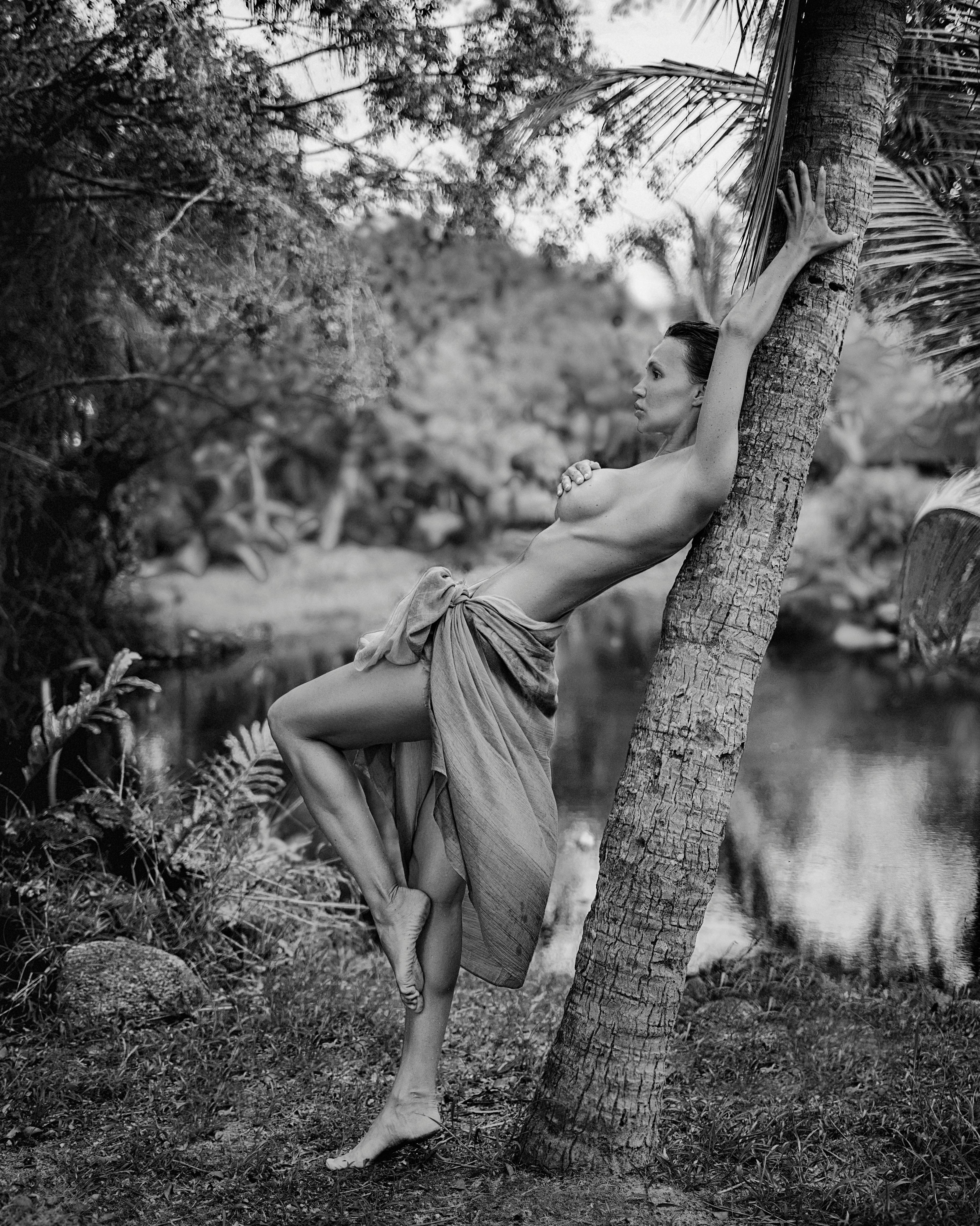 grayscale photo of a shirtless woman leaning on a tree