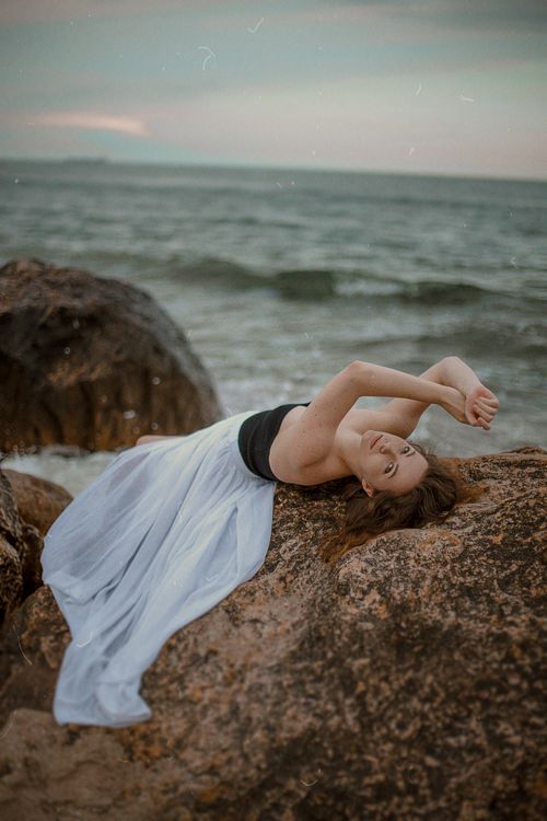 Free A woman elegantly poses on rocks by the sea, with waves in the background and a serene twilight sky. Stock Photo