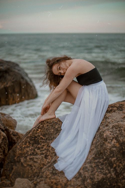 Free A woman in a flowing dress poses gracefully on beach rocks by the sea during a calm evening. Stock Photo