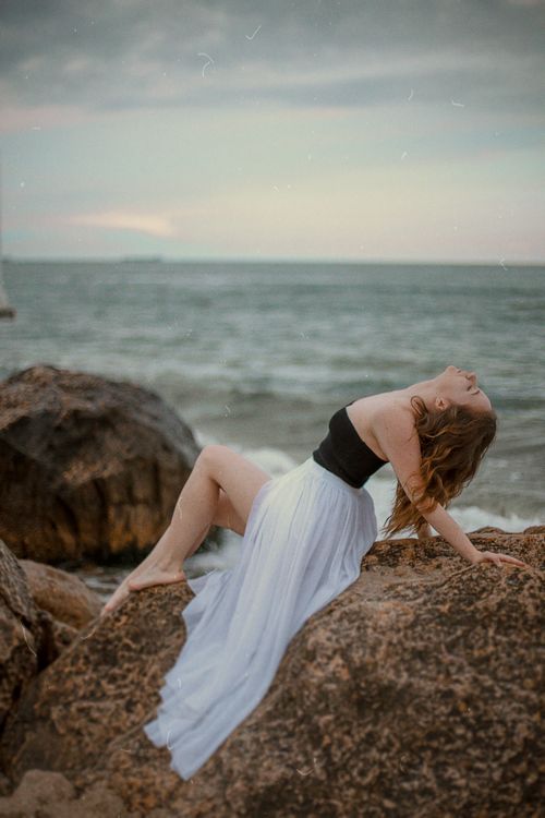 Free A woman gracefully posing on beach rocks in a white dress, with the ocean in the background. Stock Photo