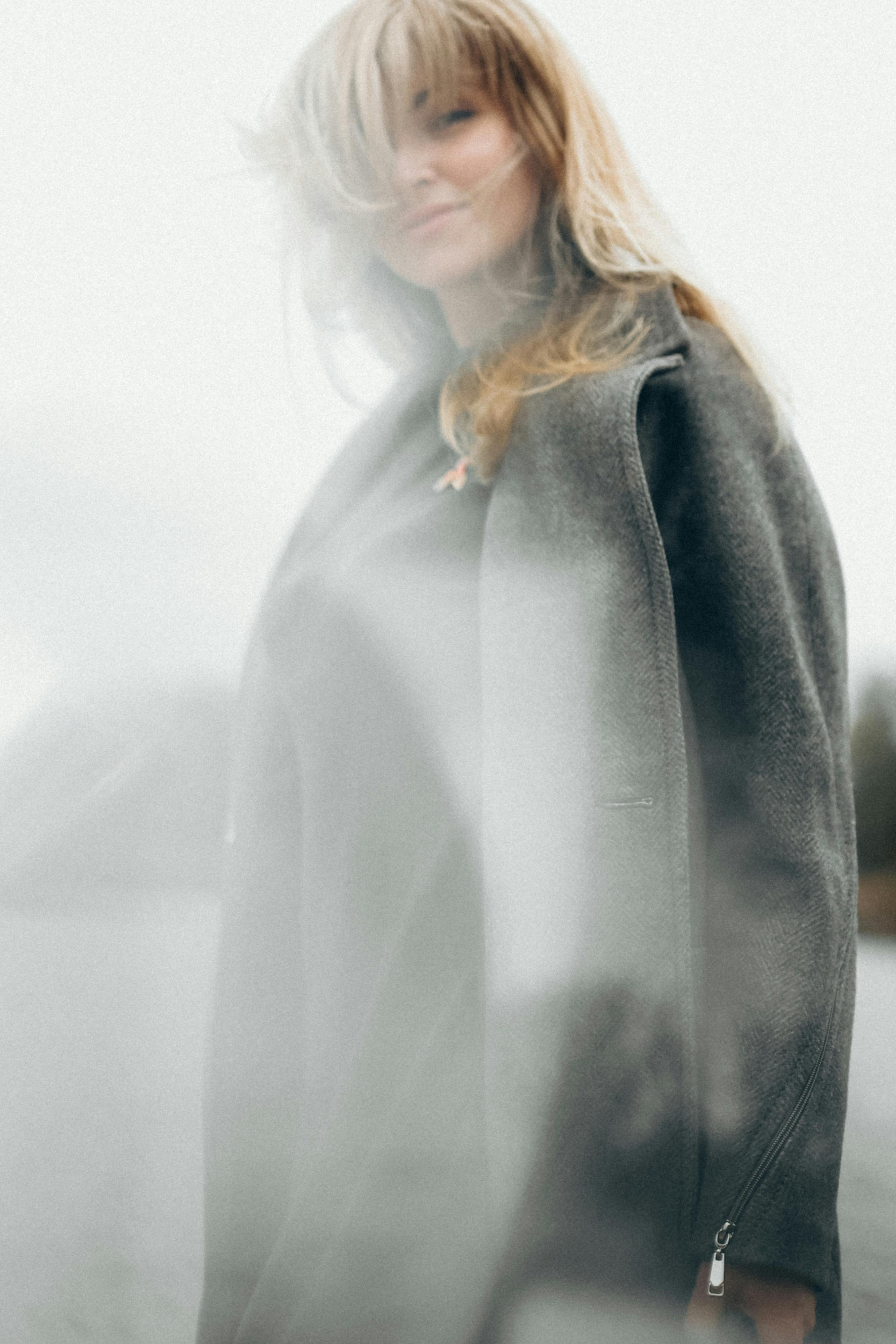 Soft-focus outdoor portrait of a woman with wind-swept hair wearing a gray coat, creating a dreamy atmosphere.
